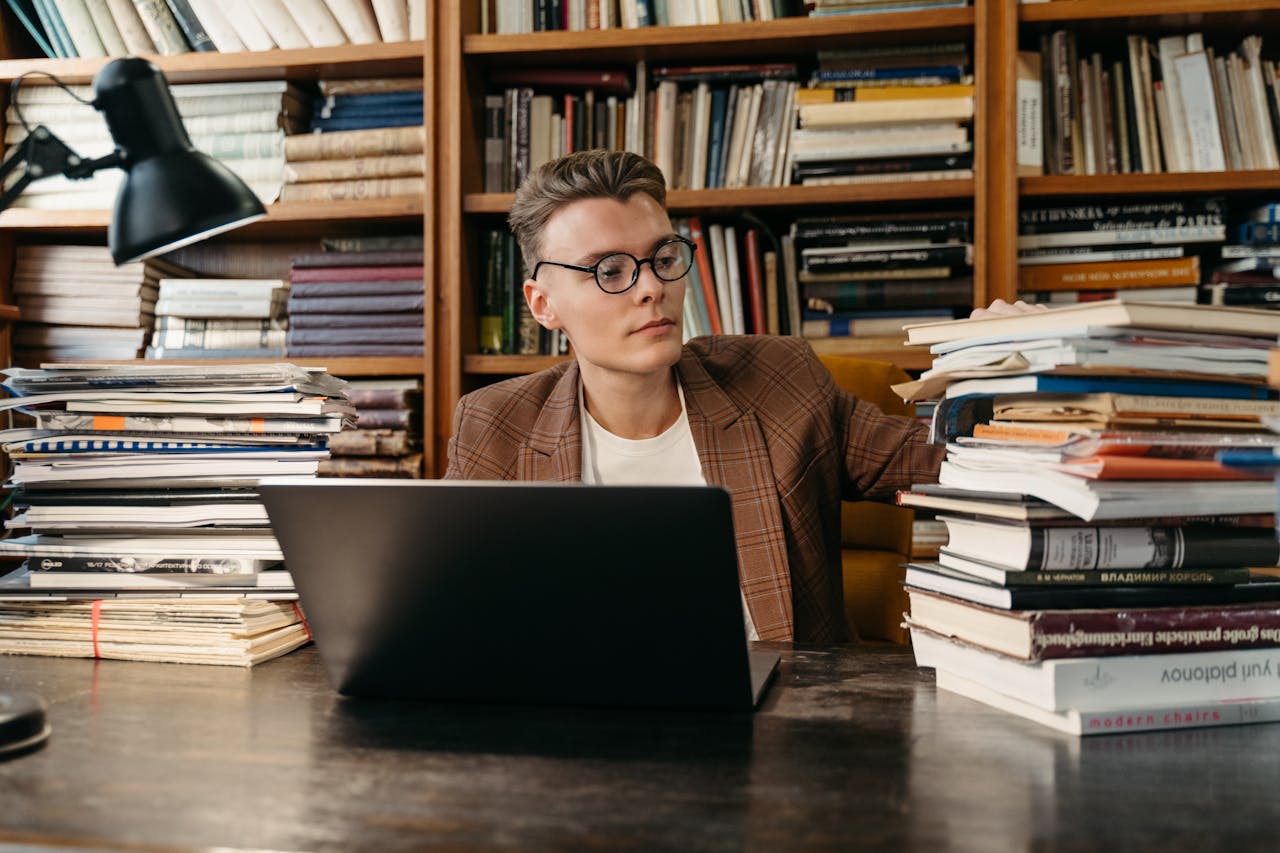 A young man with glasses working on a laptop surrounded by books in a library.